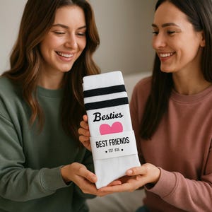 Puede incluir: Calcetines blancos con rayas negras y las palabras "Besties" y "Best Friends" impresas. Una forma de coraz&oacute;n rosa est&aacute; entre las palabras. Los calcetines son sostenidos por dos personas.