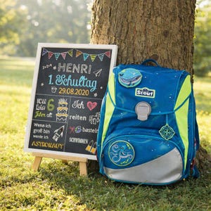Peut inclure: Un sac &agrave; dos Scout bleu et vert avec une bande r&eacute;fl&eacute;chissante argent&eacute;e, &agrave; c&ocirc;t&eacute; d'un tableau noir. Le tableau porte le nom "HENRI" et la date "29.08.2020". Le sac &agrave; dos a plusieurs &eacute;cussons, dont une baleine et une pieuvre.