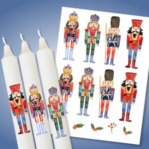 Può includere: Candele affusolate bianche e un foglio di carta con illustrazioni ad acquerello di soldati schiaccianoci in uniformi rosse, blu e verdi. Le candele hanno disegni di schiaccianoci abbinati. Lo sfondo è sfumato blu.