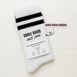 Könnte beinhalten: Weiße Socke mit schwarzen Streifen am oberen Rand. Auf der Socke steht der Text "COOLE SOCKE seit 1976". Ein Etikett auf der Socke lautet "SCHONEND BEHANDELN DAS GUTE STÜCK IST" mit Waschanweisungen. Der Text "Jedes Datum möglich" ist ebenfalls sichtbar.