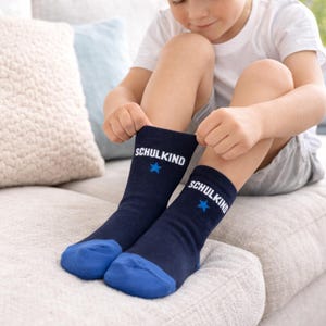 Peut inclure: Paire de chaussettes bleu marine avec des pointes bleues. Le mot "SCHULKIND" est imprim&eacute; en blanc, accompagn&eacute; d'une &eacute;toile bleue. Un enfant tire sur les chaussettes.