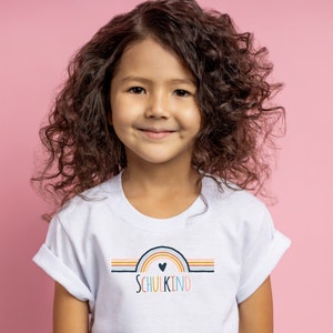 May include: A white short-sleeved t-shirt featuring a graphic of a multi-colored rainbow with a small heart at the center and the text "SCHULKIND" printed in multi-colored letters below the rainbow design, centered on the chest of the garment.