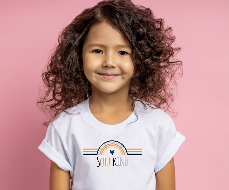 May include: A white short-sleeved t-shirt featuring a graphic of a multi-colored rainbow with a small heart at the center and the text "SCHULKIND" printed in multi-colored letters below the rainbow design, centered on the chest of the garment.