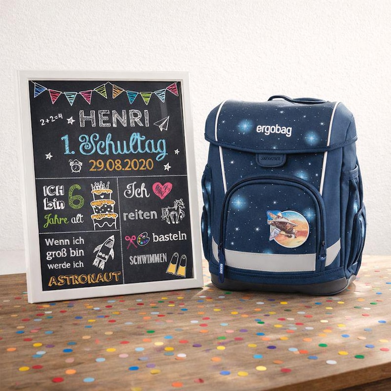Peut inclure: Un sac &agrave; dos scolaire bleu marine avec un motif sur le th&egrave;me de l'espace, avec des &eacute;toiles et une fus&eacute;e. &Agrave; c&ocirc;t&eacute;, un tableau noir avec le texte "HENRI 1. Schultag 29.08.2020" et d'autres notes et dessins manuscrits. La sc&egrave;ne se d&eacute;roule sur une table en bois.