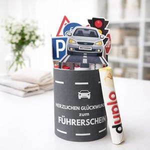 May include: A cylindrical gift box with a road design, featuring a pop-up car and road signs. The box has the German text "Herzlichen Glückwunsch zum Führerschein". A rolled-up item with the word "duplo" is next to the box.