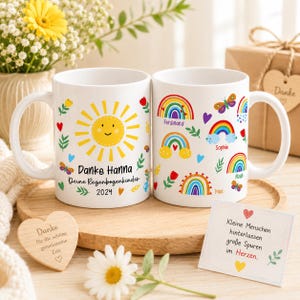 Pode incluir: Duas canecas de cerâmica branca com designs coloridos. Uma caneca apresenta um sol sorridente, o texto "Danke Hanna Deine Regenbogenkinder 2024" e elementos decorativos. A outra caneca tem arco-íris, borboletas e nomes. Um coração de madeira e um cartão também são visíveis.