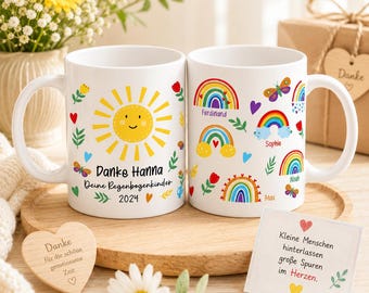 May include: Two white ceramic mugs with colorful designs. One mug features a smiling sun, the text "Danke Hanna Deine Regenbogenkinder 2024", and decorative elements. The other mug has rainbows, butterflies, and names. A wooden heart and a card are also visible.
