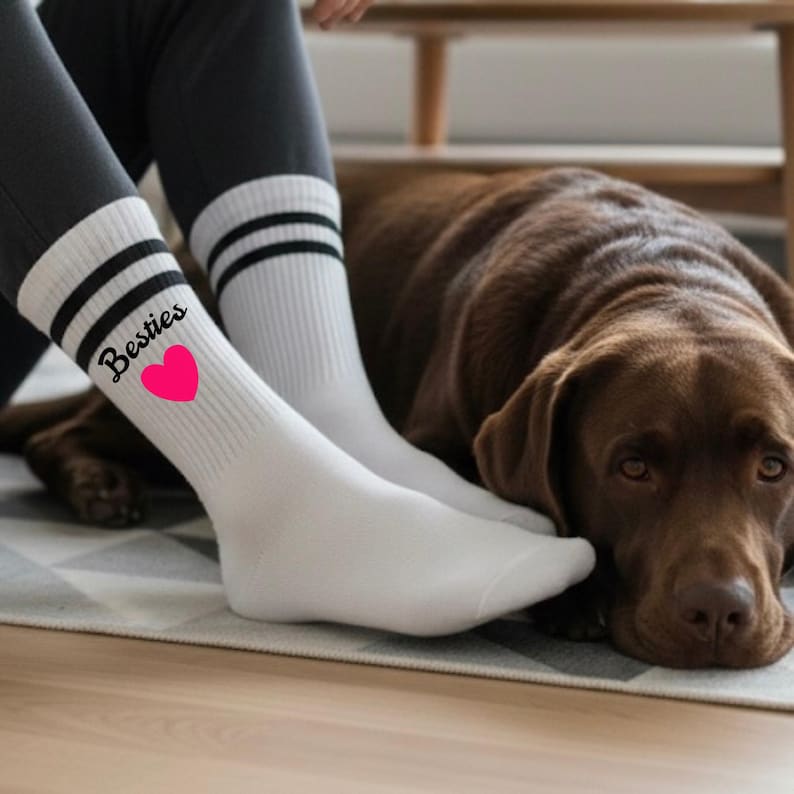 Puede incluir: Calcetines blancos con rayas negras y la palabra "Besties" en cursiva con un coraz&oacute;n rosa. Los calcetines se usan con leggings grises oscuros. Un perro marr&oacute;n est&aacute; acostado cerca.