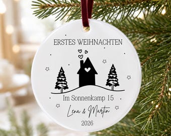 May include: A round white ceramic ornament hanging from a dark red velvet ribbon. The front features black printed graphics of a house with a heart, two pine trees, stars, and the text ERSTES WEIHNACHTEN, Im Sonnenkamp 15, Lena & Martin, 2026.