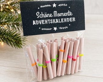 May include: A clear plastic bag containing rolled light pink paper scrolls secured with small neon-colored rubber bands. The bag is sealed with a black header card featuring white text that reads Schöne Momente ADVENTSKALENDER.