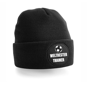 Könnte beinhalten: Schwarze Strickmütze mit einem runden Aufnäher mit einem Fußball-Grafik und den Worten "WELTBESTER TRAINER" in Weiß. Ein klassischer Stil, geeignet für Sportbegeisterte.