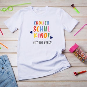 Pu&ograve; includere: Maglietta bianca con il testo "ENDLICH SCHUL KIND!" in colori arcobaleno e "HIPP HIPP HURRA!" in blu. La maglietta &egrave; su una superficie di legno con cannucce colorate e articoli per feste.