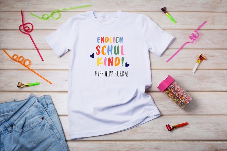 Pu&ograve; includere: Maglietta bianca con il testo "ENDLICH SCHUL KIND!" in colori arcobaleno e "HIPP HIPP HURRA!" in blu. La maglietta &egrave; su una superficie di legno con cannucce colorate e articoli per feste.
