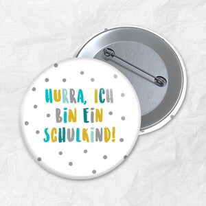 Puede incluir: Un botón blanco con lunares grises y el texto "Hurra, ich bin ein Schulkind!" en letras amarillas, azules y verdes.