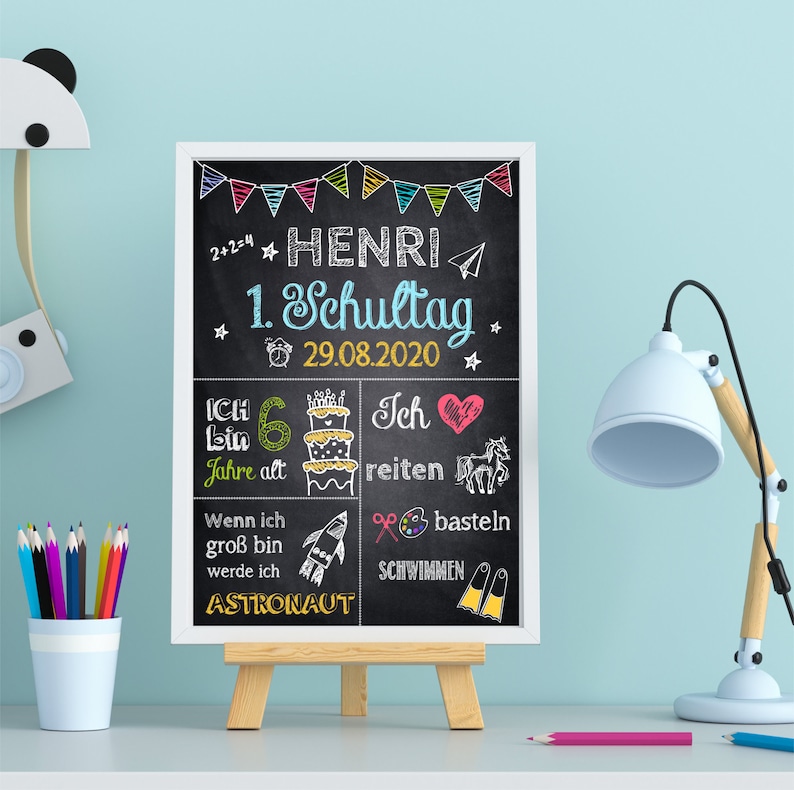 Peut inclure: Un tableau noir avec le nom "HENRI" et la date "29.08.2020" en lettres blanches. Le tableau comprend des dessins color&eacute;s et du texte en allemand, comme "1. Schultag" et "Astronaut". Le tableau est affich&eacute; sur un petit chevalet en bois.