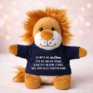 Può includere: Un leone di peluche con corpo marrone, criniera arancione e maglietta blu navy. La maglietta ha un testo bianco: "SEI MUTIG WIE ein Löwe, STEH AUF UND GEH VORAN, GLAUB FEST AN DEINE STÄRKE, WEIL JEDER ALLES SCHAFFEN KANN."