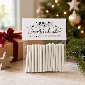 Può includere: Un sacchetto di plastica trasparente contenente ventiquattro rotolini di carta bianca, sormontato da un cartoncino bianco con illustrazioni nere di tre gnomi e il testo Adventskalender 24 LOSE WEIL DU MIR WICHTELIG BIST.