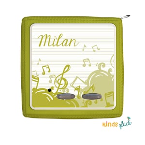 Op de afbeelding: Een groene stoffen muziekspeler met een wit gezicht en de naam "Milan" erop gedrukt. Het gezicht is versierd met muzieknoten en een solsleutel.