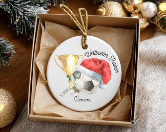 Op de afbeelding: Een witte keramische kerstversiering met een voetbal met een kerstmuts, een gouden trofee en de tekst "Weltbester Trainer" en "Clemens". De versiering zit in een bruine geschenkdoos met een gouden touwtje.