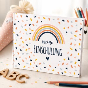 Könnte beinhalten: Ein rechteckiges Buch mit weißem Einband, verziert mit einer bunten Regenbogen-Grafik, verstreuten Konfetti-Mustern in Orange, Blau und Gelb sowie dem Schriftzug "meine EINSCHULUNG" in dunkelblauer Schreib- und Druckschrift mit einem Herz.