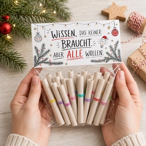 Può includere: Un sacchetto di plastica trasparente contenente diversi rotoli di carta fissati con piccoli elastici colorati. L'intestazione in cartoncino riporta il testo nero e rosso WISSEN, DAS KEINER BRAUCHT. ABER ALLE WOLLEN. con illustrazioni di decorazioni natalizie.