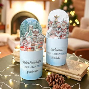 Könnte beinhalten: Zwei Weihnachtskarten zum Aufklappen. Eine zeigt ein Winterdorf mit Schneemännern und Geschenken. Die andere zeigt den Weihnachtsmann, einen Pinguin und ein Auto. Auf den Karten steht "Kleine Botschaften vom Nikolaus persönlich" und "Eine Portion Weihnachtszauber für dich."