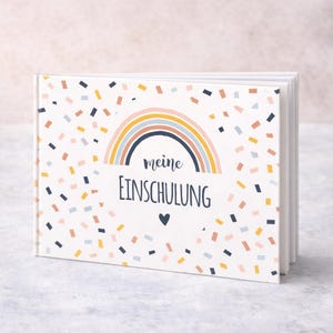 May include: A white hardcover book featuring a colorful rainbow graphic and the text "meine EINSCHULUNG" with a small heart icon. The cover is decorated with a pattern of small, scattered rectangular confetti shapes in shades of blue, yellow, and pink.