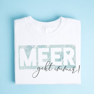 Op de afbeelding: Wit T-shirt met een blauwe en witte afbeelding met de tekst "MEER...geht immer!" met een klein hartje in de "R".