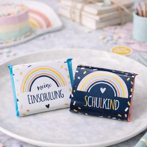 May include: Two rectangular chocolate bars wrapped in paper. The left wrapper is white with a pastel rainbow, confetti, and the text 'meine EINSCHULUNG'. The right wrapper is dark blue with a pastel rainbow, confetti, and the text 'SCHULKIND'.