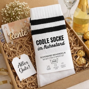 Könnte beinhalten: Geschenkbox mit weißen Socken mit schwarzen Streifen und dem Text "COOLE SOCKE im Ruhestand". Die Box enthält auch ein Etikett mit "Alles Gute!" und ein Holzschild mit "Rente".