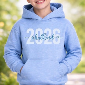 Peut inclure: Un sweat &agrave; capuche bleu clair avec une poche avant. Le sweat-shirt affiche le texte "2026 schulkind" en blanc et bleu clair. Le mot "schulkind" est &eacute;crit en cursive avec un petit symbole de c&oelig;ur.