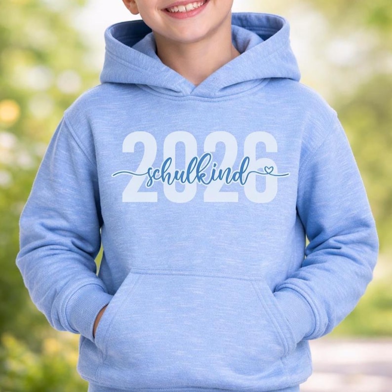 Peut inclure: Un sweat &agrave; capuche bleu clair avec une poche avant. Le sweat-shirt affiche le texte "2026 schulkind" en blanc et bleu clair. Le mot "schulkind" est &eacute;crit en cursive avec un petit symbole de c&oelig;ur.