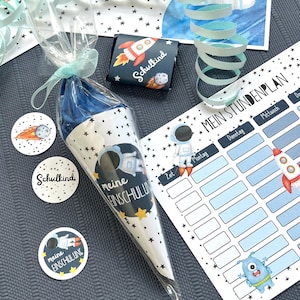 Puede incluir: Un conjunto de útiles escolares con temática espacial. Incluye una bolsa de regalo en forma de cono con un diseño de astronauta y estrellas, pegatinas, una barra de chocolate y un horario semanal. El texto en la bolsa dice "meine Einschulung".