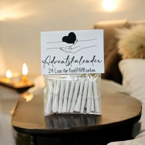 Puede incluir: Una bolsa transparente llena de 24 papeles blancos enrollados, cada uno asegurado con una banda blanca. La bolsa está coronada con una tarjeta blanca con un corazón negro y las palabras "Adventskalender" y "24 Lose für KostPAARzeiten" en escritura negra.