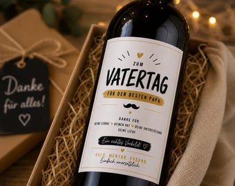 May include: A dark wine bottle with a white label that reads "ZUM VATERTAG" and "FÜR DEN BESTEN PAPA". The bottle is in a gift box with a tag that says "Danke für alles!" and a heart symbol.