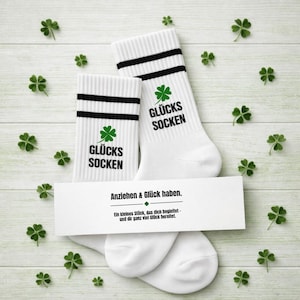 Könnte beinhalten: Weiße Socken mit schwarzen Streifen und dem Text "GLÜCKS SOCKEN" und einem grünen Kleeblatt. Eine weiße Karte mit dem Text "Anziehen & Glück haben" und einer Nachricht ist darunter platziert. Grüne Kleeblätter sind verstreut.