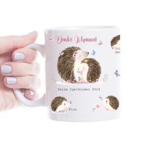 Op de afbeelding: Witte keramische mok met illustraties van egels en vlinders. De mok heeft de tekst "Danke Johanna!" en "Deine Igelkinder 2024", evenals namen. De mok heeft een wit handvat.