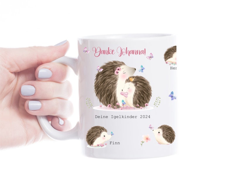 Op de afbeelding: Witte keramische mok met illustraties van egels en vlinders. De mok heeft de tekst "Danke Johanna!" en "Deine Igelkinder 2024", evenals namen. De mok heeft een wit handvat.