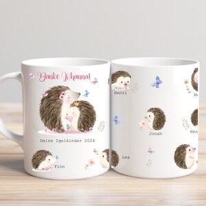 Op de afbeelding: Witte keramische mok met een aquarel illustratie van twee egels met de tekst "Danke Johanna!" en "Deine Igelkinder 2024". De mok heeft ook kleinere egel illustraties met namen en vlinders.