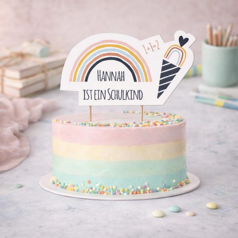 May include: A round cake with pastel horizontal stripes of pink, yellow, and blue, topped with colorful sprinkles. A white cake topper features a rainbow, an ice cream cone, the text 1+1=2, and the name Hannah with the text IST EIN SCHULKIND.