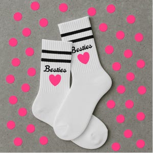 Puede incluir: Un par de calcetines blancos con rayas negras y la palabra "Besties" impresa en negro, junto con un coraz&oacute;n rosa. Los calcetines se muestran sobre una superficie gris con lunares rosas dispersos.