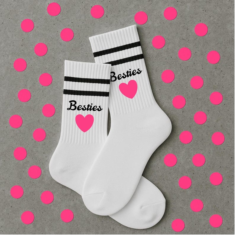 Puede incluir: Un par de calcetines blancos con rayas negras y la palabra "Besties" impresa en negro, junto con un coraz&oacute;n rosa. Los calcetines se muestran sobre una superficie gris con lunares rosas dispersos.