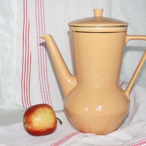 Könnte beinhalten: Eine pfirsichfarbene Kaffeekanne aus Keramik mit Deckel und langem Ausguss. Die Kanne hat einen Griff und steht auf einem weißen Tuch mit roten Streifen. Ein rot-gelber Apfel liegt links von der Kanne.