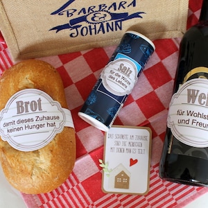 Könnte beinhalten: Ein rot-weiß kariertes Tischtuch mit einem Laib Brot, einer Flasche Wein, einem Salzstreuer und einer Karte mit dem Text "DAS SCHONSTE AM ZUHAUSE SIND DIE MENSCHEN MIT DENEN MAN ES TEILT". Das Brot hat ein Etikett mit der Aufschrift "Brot damit dieses Zuhause keinen Hunger hat". Die Weinflasche hat ein Etikett mit der Aufschrift "Wein für Wohlstand und Freunde". Der Salzstreuer hat ein Etikett mit der Aufschrift "Salz für die Würze des Lebens".