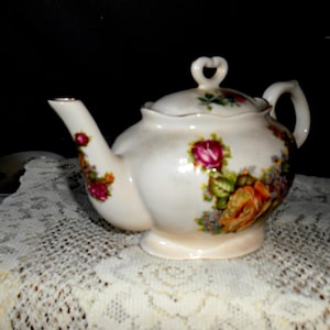 May include: Cream-colored ceramic teapot with a floral design featuring pink and orange roses, green leaves, and blue accents. The teapot has a heart-shaped handle and a matching lid. A gold trim accents the spout and lid. The teapot is sitting on a white lace tablecloth.