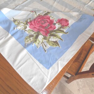 May include: A square, vintage tablecloth featuring a large floral design. The central motif showcases a cluster of red roses with green leaves, set against a light blue and white striped background. The tablecloth is folded on a wooden surface.