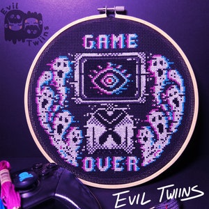 May include: A black and white cross-stitch embroidery hoop with the words "GAME OVER" and a pixelated eye design. The hoop is surrounded by a wooden frame and the words "Evil Twins" are written below the hoop.
