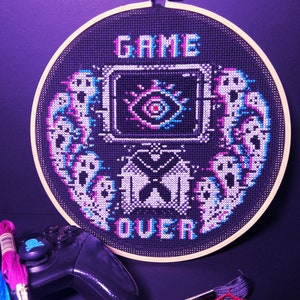 May include: A black and white cross-stitch embroidery hoop with the text "GAME OVER" in a pixelated font. The design features a pixelated eye with a red pupil and a pixelated butterfly. The embroidery is framed in a wooden hoop.