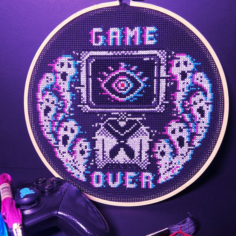 May include: A black and white cross-stitch embroidery hoop with the text "GAME OVER" in a pixelated font. The design features a pixelated eye with a red pupil and a pixelated butterfly. The embroidery is framed in a wooden hoop.