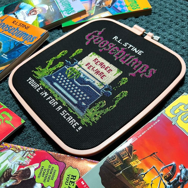 May include: A black cross-stitch featuring the text "Goosebumps" in pink and green, with a typewriter and the words "Reader Beware!" and "You're in for a scare!" surrounded by green splatters. The cross-stitch is in a pink hoop, with several Goosebumps books surrounding it.